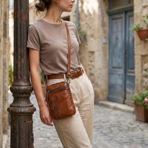 May include: A tan leather crossbody bag with a vertical zippered front pocket and a matching tan leather belt. The bag features a long adjustable strap and metal hardware, worn over a plain taupe t-shirt and light-coloured linen trousers.