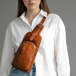 Op de afbeelding: Een bruine leren sling bag met een voorvak met ritssluiting en een verstelbare riem. De tas wordt over het lichaam gedragen en heeft een messing gesp. Het leer heeft een getextureerde uitstraling.