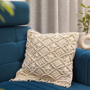 May include: A square throw pillow featuring an off-white macrame cover with a complex diamond and knot pattern. The front surface displays a textured, woven design with fringe detailing along the bottom edge, resting on a deep blue upholstered surface.