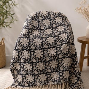 May include: A rectangular throw blanket featuring a repeating floral pattern in dark navy blue and cream. The fabric is adorned with detailed flower motifs and leaves throughout, finished with a fringe of cream-colored tassels along the bottom edge.