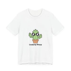 Peut inclure: T-shirt blanc avec un cactus de dessin animé portant des lunettes de soleil dans un pot en terre cuite. L'expression "Looking Sharp" est imprimée en dessous du cactus. Le motif est centré sur le t-shirt.