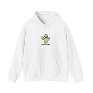 Peut inclure: Sweat à capuche blanc avec une poche avant et un cordon de serrage. Le motif représente un cactus de dessin animé portant des lunettes de soleil dans un pot, avec le texte "Looking Sharp" en dessous.