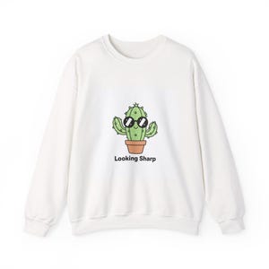 Peut inclure: Sweat-shirt blanc avec un cactus de dessin animé portant des lunettes de soleil. Le cactus vert est dans un pot marron. Le texte "Looking Sharp" est imprimé en dessous. Design décontracté et confortable.