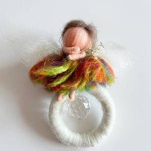 Puede incluir: Una figura de lana fieltrada con cara color melocotón y cabello castaño, que lleva una falda arcoíris multicolor y alas blancas. La figura sostiene una pequeña ramita dorada y se asienta sobre un anillo blanco envuelto en hilo con una cuenta de cristal.