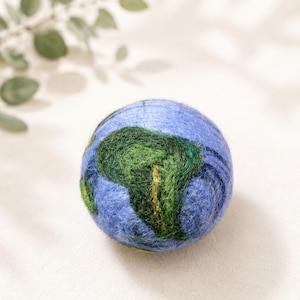 Könnte beinhalten: Ein kugelförmiger, nadelgefilzter Wollball mit einem texturierten Design der Erde. Die Oberfläche zeigt Schattierungen von Hellblau für die Ozeane und tiefes Grün mit subtilen gelben Akzenten für die Kontinente in einer flauschigen Optik.