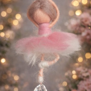 Può includere: Ornamento fatto a mano a forma di ballerina. La bambola ha un tutù rosa e un ciondolo di cristallo. La ballerina ha i capelli castani e le braccia alzate. Lo sfondo è sfocato con luci bokeh e fiori rosa.