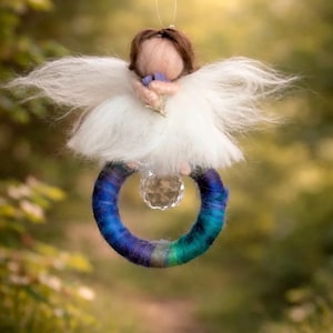Könnte beinhalten: Ein gefilzter Engel-Anhänger mit braunem Haar und weißen, flauschigen Flügeln, der einen kleinen Gegenstand hält. Die Figur sitzt auf einem kreisförmigen Ring, der mit blau, violett und türkis verlaufender Wolle umwickelt ist und eine Kristallperle.