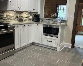 Può includere: Una cucina moderna con mobili bianchi, un forno in acciaio inossidabile e un microonde. Il paraschizzi presenta un motivo a mosaico. Il pavimento è piastrellato con grandi piastrelle rettangolari grigie. Una finestra offre una vista su un interno in legno.