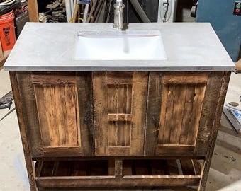 Può includere: Mobile bagno rustico in legno con piano grigio e lavabo rettangolare bianco. Il mobile ha tre ante e un ripiano inferiore. Il legno ha una finitura naturale e invecchiata. Il rubinetto è argentato.