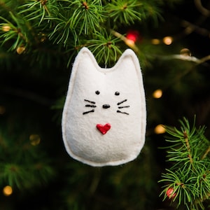 White felt cat hangs in Christmas tree branches with twinkle lights in background. Whimsical rounded shape with pointed ears. Painted black nose, eyes and whiskers, and small red painted heart near bottom.