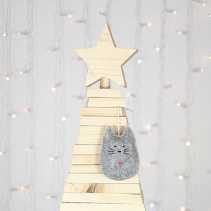 Grey felt cat ornament with hand painted black eyes, nose, whiskers and a small red heart displayed on a wooden tree with white lights in background.