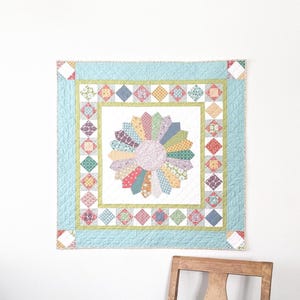 May include: A colourful quilt with a circular design in the centre, surrounded by a border of green and blue squares. The quilt is hanging on a white wall above a wooden chair.