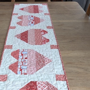 May include: A white table runner with a red and pink patchwork design featuring five hearts. The hearts are made of different fabrics with various patterns. The runner has a white background with a subtle pattern and a red border.