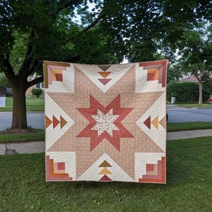 May include: A handmade quilt with a starburst design. The quilt features a central star in shades of red and cream, surrounded by geometric shapes in various earth tones. The quilt is displayed outdoors, hanging from a tree.