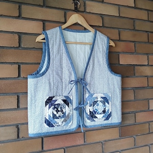 May include: A quilted vest featuring a light blue floral pattern and denim blue trim. The vest has two square pockets with a geometric patchwork design in shades of blue. It hangs on a wooden hanger against a brick wall.