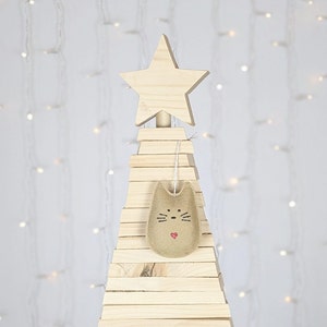 Light brown felt cat ornament with hand painted black eyes, nose, whiskers and a small red heart displayed on a wooden tree with white lights in background.