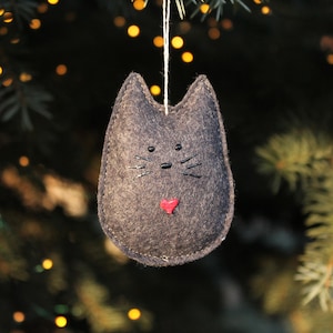 Charcoal black felt cat hangs in Christmas tree branches with twinkle lights in background. Whimsical rounded shape with pointed ears. Painted black nose, eyes and whiskers, and small red painted heart near bottom.