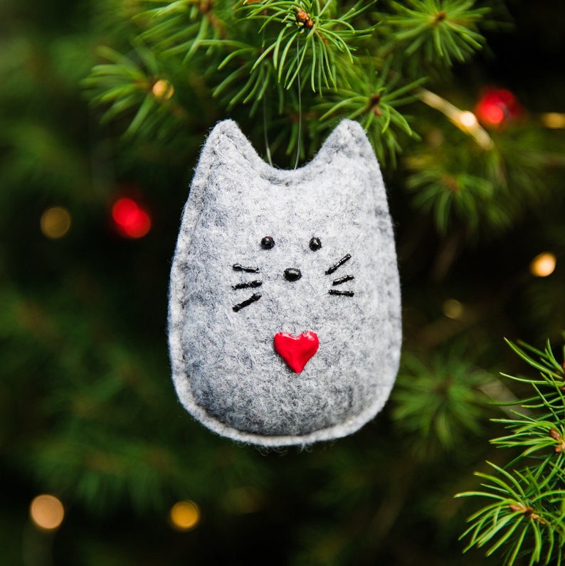 Light gray felt cat hangs in Christmas tree branches with twinkle lights in background. Whimsical rounded shape with pointed ears. Painted black nose, eyes and whiskers, and small red painted heart near bottom.