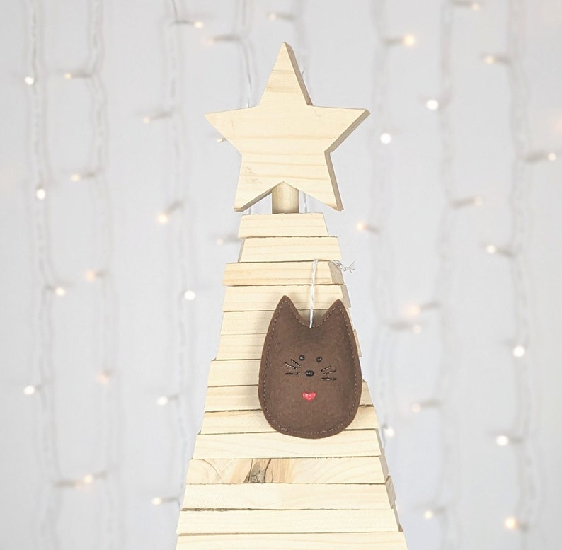 Dark brown felt cat ornament with hand painted black eyes, nose, whiskers and a small red heart and displayed on a wooden tree with white lights in background.