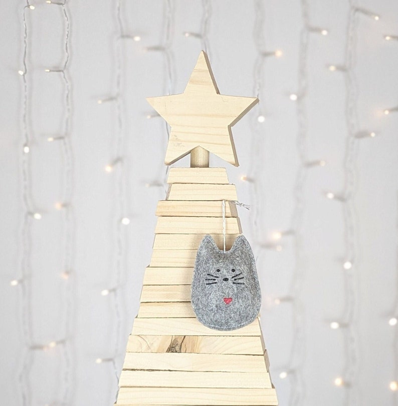 Grey felt cat ornament with hand painted black eyes, nose, whiskers and a small red heart displayed on a wooden tree with white lights in background.