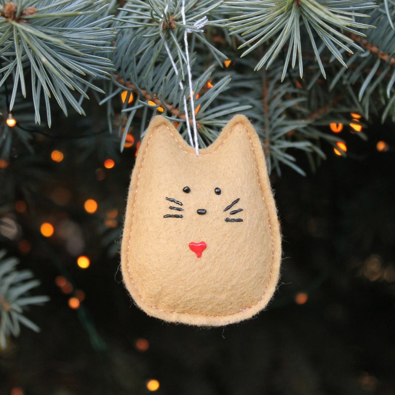 Ginger coloured felt cat hangs in Christmas tree branches with twinkle lights in background. Whimsical rounded shape with pointed ears. Painted black nose, eyes and whiskers, and small red painted heart near bottom.