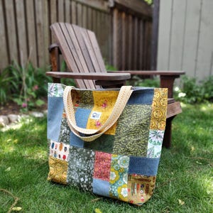 May include: A patchwork tote bag constructed from various fabric panels featuring floral, sun, and botanical patterns in shades of yellow, green, and blue. The bag includes two beige herringbone-patterned straps and a small white rectangular brand label.