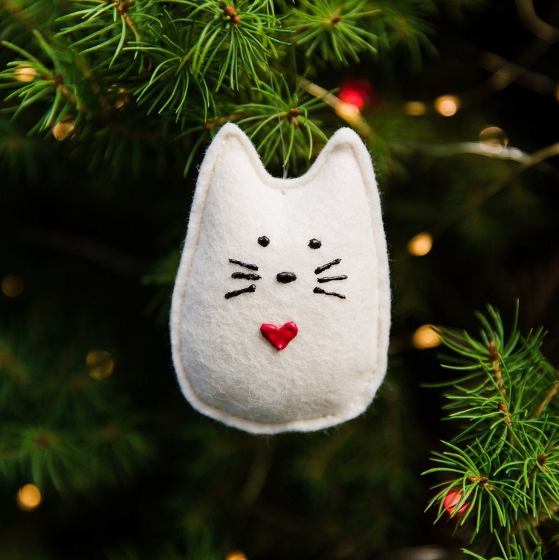 White felt cat hangs in Christmas tree branches with twinkle lights in background. Whimsical rounded shape with pointed ears. Painted black nose, eyes and whiskers, and small red painted heart near bottom.