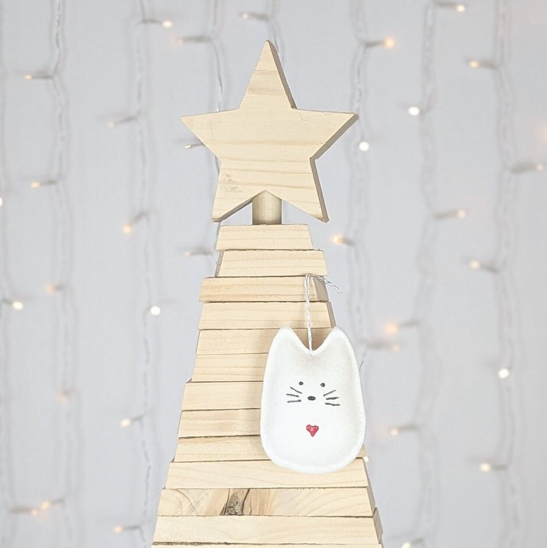 White felt cat ornament with hand painted black eyes, nose, whiskers and a small red heart displayed on a wooden tree with white lights in background.