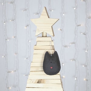 Black felt cat ornament with hand painted black eyes, nose, whiskers and a small red heart displayed on a wooden tree with white lights in background.