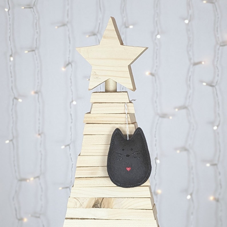 Black felt cat ornament with hand painted black eyes, nose, whiskers and a small red heart displayed on a wooden tree with white lights in background.