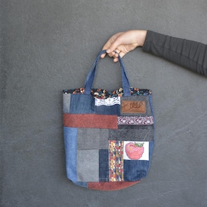 Puede incluir: Un bolso tote patchwork con cuadrados de tela vaquera azul, roja y gris. El bolso tiene asas azules y un pequeño parche con un diseño de manzana. Una etiqueta de cuero marrón con la palabra "ROSA" está cosida en la parte superior.