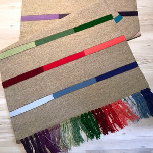 Puede incluir: Dos alfombras tejidas con flecos coloridos. La alfombra superior tiene rayas verdes, moradas y verdes. La alfombra inferior tiene rayas verdes, rojas, azules y blancas.