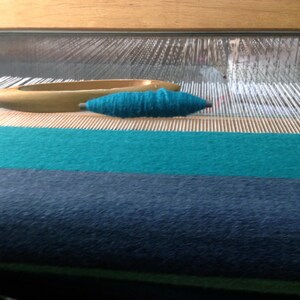 May include: A close-up of a loom with a teal colored yarn wound on a shuttle. The loom is set up with teal and dark blue fabric already woven.