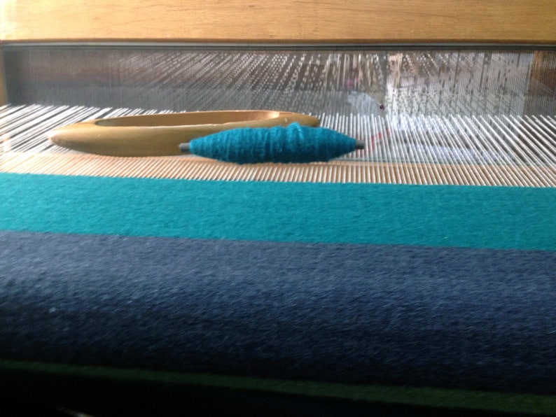 May include: A close-up of a loom with a teal colored yarn wound on a shuttle. The loom is set up with teal and dark blue fabric already woven.