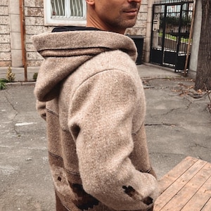 May include: A hooded jacket in shades of brown and tan. The jacket has a textured, woven appearance with darker brown accents and a contrasting lower panel. The person is wearing blue jeans.