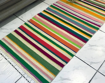 Puede incluir: Una alfombra rayada colorida con flecos blancos. La alfombra está hecha de lana y tiene una variedad de colores, incluyendo rojo, verde, amarillo y azul.