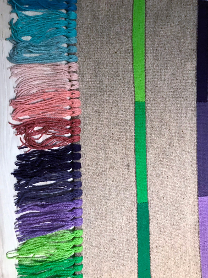 Puede incluir: Una alfombra tejida con un borde de flecos en varios colores, incluyendo azul, rosa, rojo, morado y verde. La alfombra tiene una franja verde en el centro y una franja morada en el lado derecho.