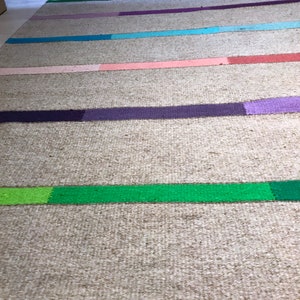 Puede incluir: Una alfombra tejida con un fondo beige natural y rayas de colores en tonos de rosa, morado, verde, azul y rojo. La alfombra tiene un borde de flecos.