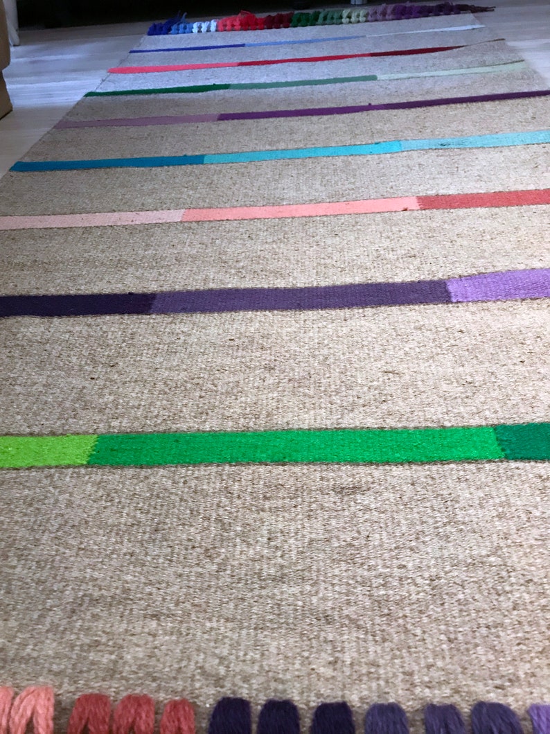 Puede incluir: Una alfombra tejida con un fondo beige natural y rayas de colores en tonos de rosa, morado, verde, azul y rojo. La alfombra tiene un borde de flecos.