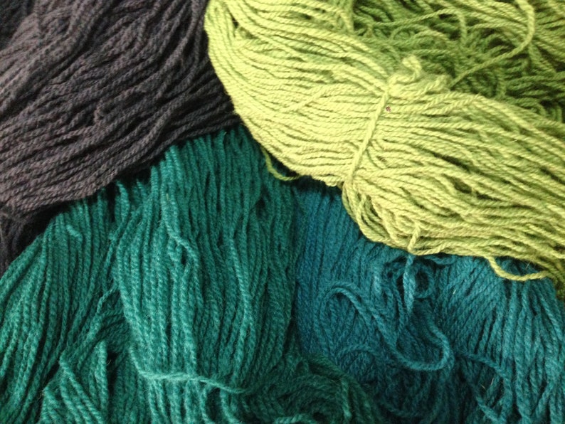 May include: A collection of yarn in shades of green, teal, and black. The yarn is arranged in a pile, with the green yarn on top.