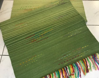 May include: Two green woven rugs with colorful fringe. The rugs have a striped pattern and are in shades of green. The fringe includes red, yellow, blue, and other colors. The rugs are rectangular and appear to be handmade.
