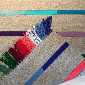 Puede incluir: Una alfombra tejida con un borde de flecos en tonos de verde, rojo, blanco, azul y morado. La alfombra tiene un fondo gris claro con rayas de colores.