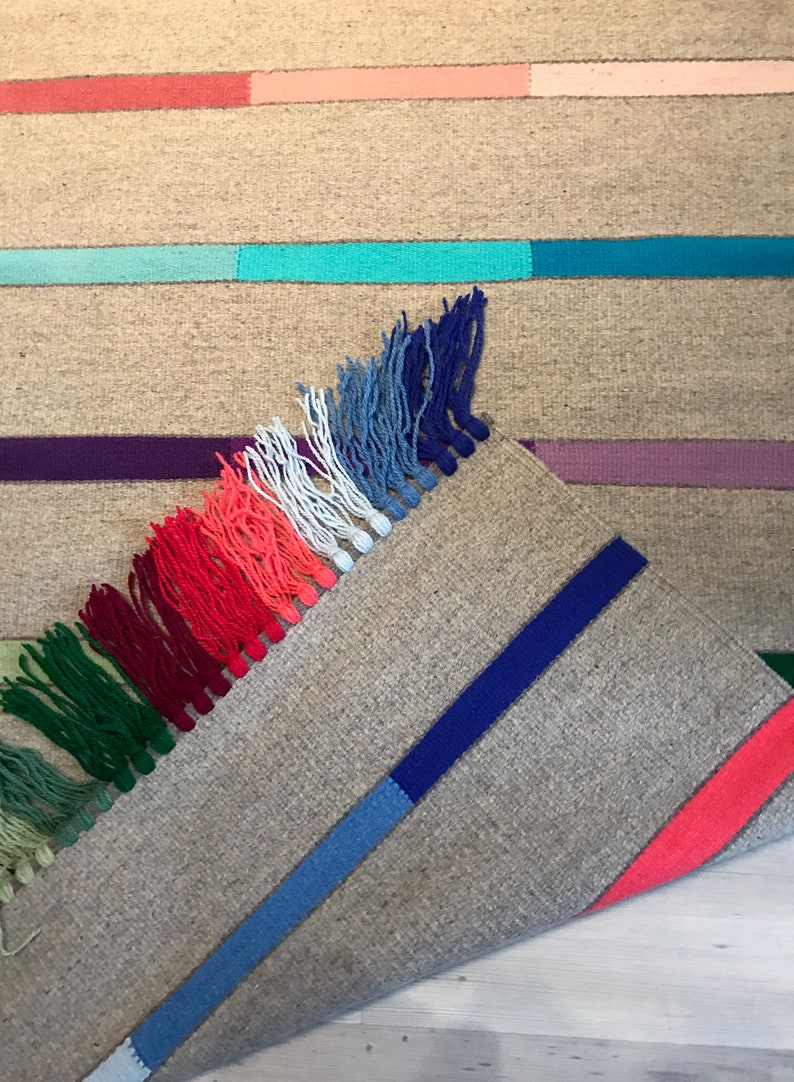 Puede incluir: Una alfombra tejida con un borde de flecos en tonos de verde, rojo, blanco, azul y morado. La alfombra tiene un fondo gris claro con rayas de colores.