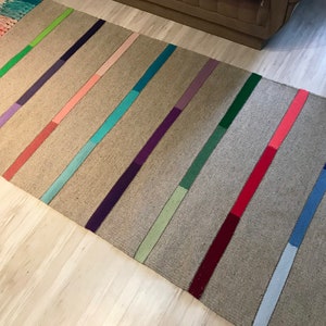 Puede incluir: Una alfombra rectangular larga con fondo gris y rayas de colores. Las rayas son de varios tonos de verde, morado, rosa, azul y rojo.