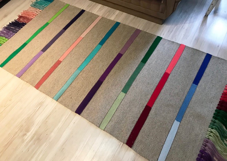 Puede incluir: Una alfombra rectangular larga con fondo gris y rayas de colores. Las rayas son de varios tonos de verde, morado, rosa, azul y rojo.