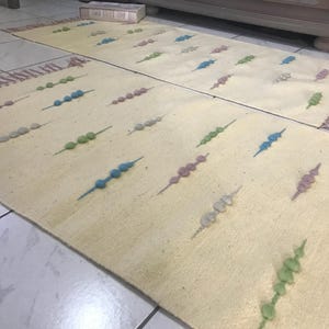 May include: Two rectangular, cream-colored rugs with colorful beaded designs. The rugs have a fringe on one end and are decorated with rows of blue, green, pink, and gray beads. The rugs are placed on a tiled floor.