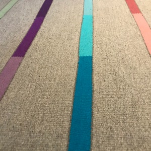 Puede incluir: Primer plano de una alfombra tejida con rayas verticales de diferentes colores, incluyendo morado, azul, azul verdoso, rosa y verde. La alfombra tiene un fondo beige natural.