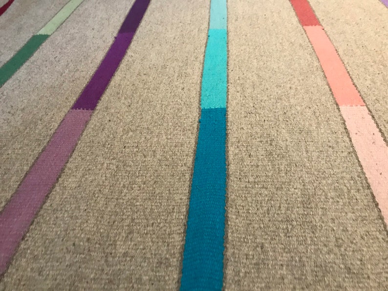 Puede incluir: Primer plano de una alfombra tejida con rayas verticales de diferentes colores, incluyendo morado, azul, azul verdoso, rosa y verde. La alfombra tiene un fondo beige natural.