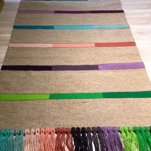 Puede incluir: Una alfombra rectangular larga con un fondo beige natural y rayas de colores. Las rayas son en tonos de azul, verde, rojo, rosa y morado. La alfombra tiene flecos en el borde inferior.