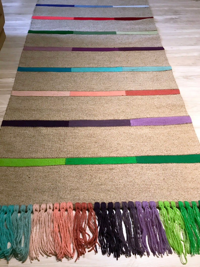 Puede incluir: Una alfombra rectangular larga con un fondo beige natural y rayas de colores. Las rayas son en tonos de azul, verde, rojo, rosa y morado. La alfombra tiene flecos en el borde inferior.